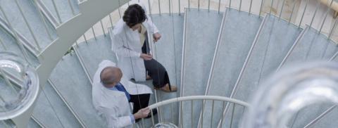 Workers wearing lab coats are engaged in conversation as they walk up a staircase.